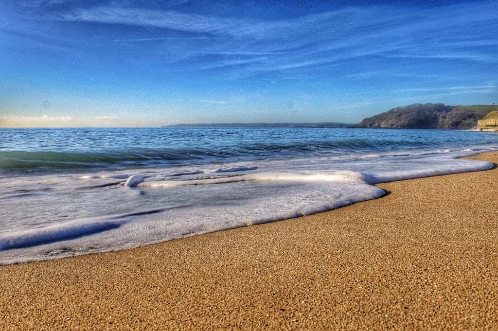 Serene view of a sandy beach in Cornwall, England, with gentle ocean waves and a clear blue sky.